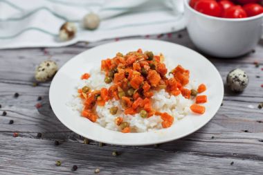 haşlanmış havuç, bezelye ve soğanlı beyaz tabakta, bıldırcın yumurtası ile lezzetli pirinç kase kiraz domates, biber ve açık gri renkli bir ışık ahşap masada havlu ile