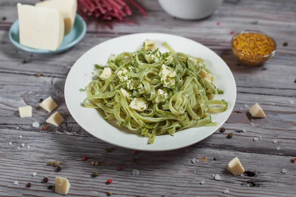 Pâtes vertes aux épinards et fromage mozzarella et légumes verts dans une assiette blanche, moutarde française, spaghettis crus colorés, soucoupe au fromage, aneth et bol aux tomates cerises sur une table en bois léger — Photo