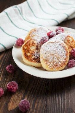 Beyaz bir tabak, bir elek, havlu ve koyu ahşap bir masa üzerinde Dondurulmuş çilek pudra şekeri ile syrniki