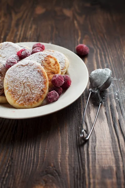 Syrniki with powdered sugar on a white plate, sieve and frozen berries on a dark wooden table — Stock Photo, Image