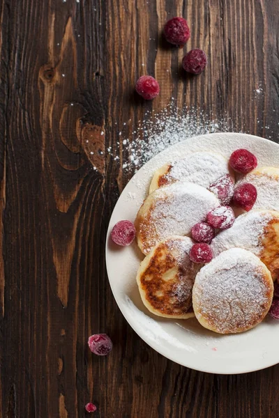 Syrniki dengan bubuk gula di piring putih, saringan dan buah beri beku di atas meja kayu yang gelap — Stok Foto