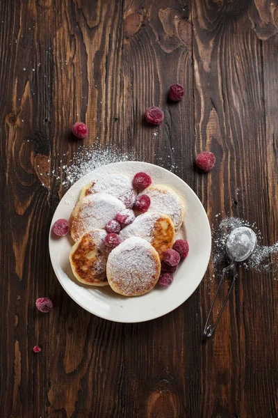 Syrniki mit Puderzucker auf einem weißen Teller, Sieb und gefrorenen Beeren auf einem dunklen Holztisch — Stockfoto