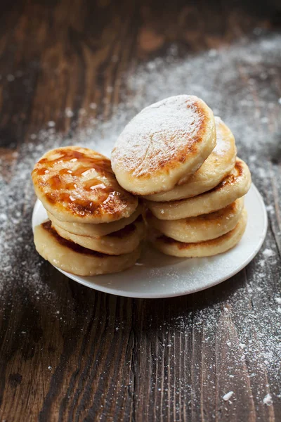 syrniki with powdered sugar on a white plate, a sieve and caramel topping on a dark wooden table