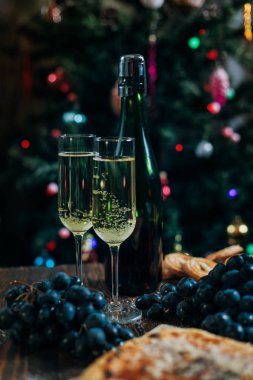 bir yeni yıl'ın tablo, peynir pasta çikolata, mandalina, üzüm, bisküvi, şampanya ve iki bardak dekore edilmiş bir Noel ağacı ışıkları ile fon karşı