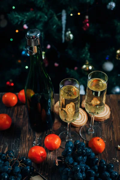Un festoso tavolo di Capodanno, torta al formaggio con cioccolato, mandarini, uva, biscotti, champagne e due bicchieri sullo sfondo di un albero di Natale decorato con luci — Foto Stock