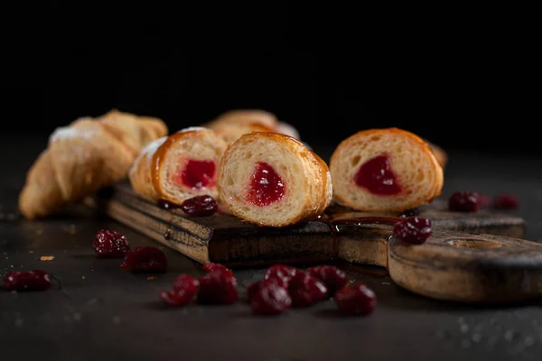 cut croissants with red berry stuffing, sprinkled with powdered sugar and poured with topping and jam, a dark cutting wooden board and red berries on a dark background