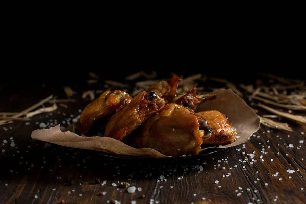 Pollo jugoso al horno con una corteza crujiente, en especias, en un plato negro, sobre papel de hornear, sal marina grande y virutas de madera grandes sobre un fondo de madera oscura — Foto de Stock