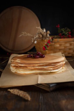 bir yığın krep, spikelets, yuvarlak kesme tahtası, dalları kırmızı meyveler ve koyu ahşap bir masa üzerinde meyveleri ile sepet