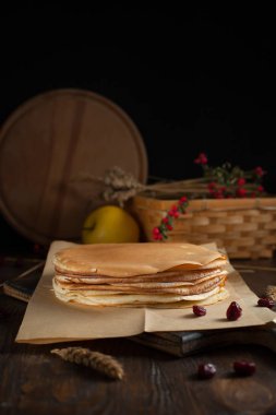 bir yığın krep, spikelets, yuvarlak kesme tahtası, dalları kırmızı meyveler ve koyu ahşap bir masa üzerinde meyveleri ile sepet