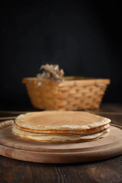 Uma pilha de panquecas, espiguetas, uma tábua de corte redonda, uma cesta com ramos com bagas vermelhas e bagas em uma mesa de madeira escura — Fotografia de Stock