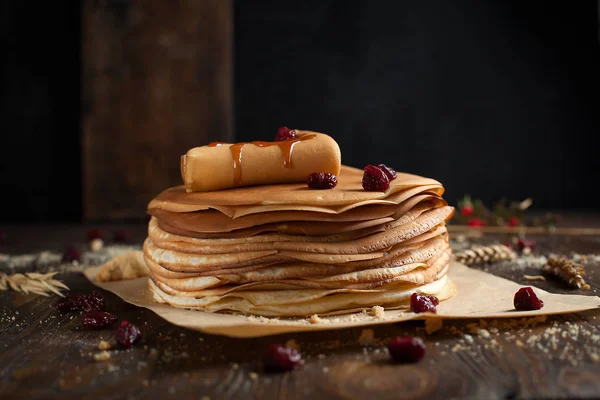 Stack of pancakes, folded pancake, sprinkled with berries, cookies, sugar powder and caramel syrup on parchment on a dark wooden table — Stock Photo, Image