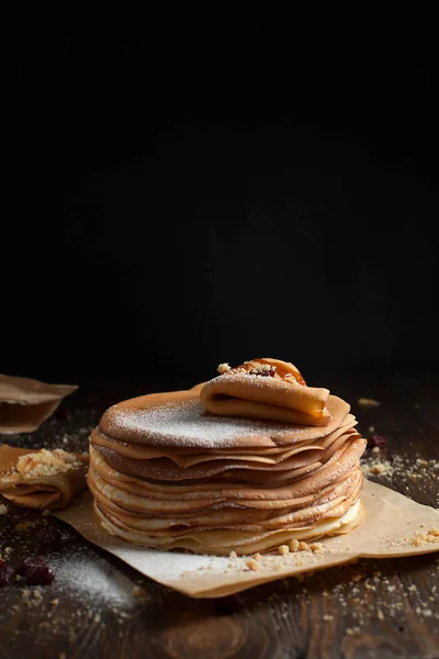Pila de panqueques, panqueque plegado, espolvoreado con bayas, galletas, azúcar en polvo y jarabe de caramelo en el pergamino sobre una mesa de madera oscura — Foto de Stock