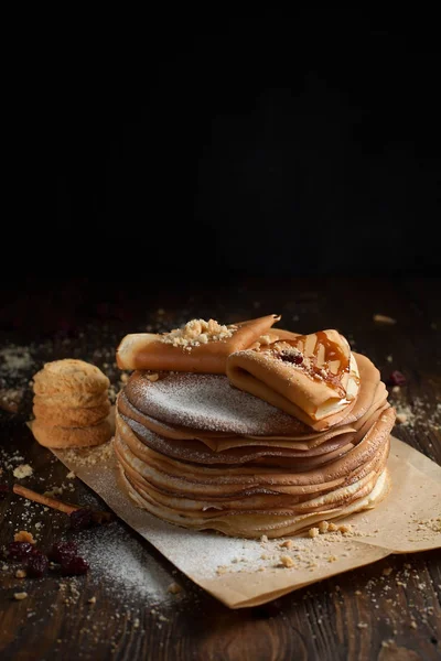 Una pila de panqueques, dos panqueques plegados, espolvoreados con bayas, galletas, azúcar en polvo y jarabe de caramelo en pergamino sobre una mesa de madera oscura — Foto de Stock