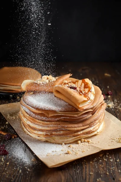 Dos pilas de panqueques, dos panqueques plegados, espolvoreados con bayas, galletas, azúcar en polvo y jarabe de caramelo en pergamino sobre una mesa de madera oscura — Foto de Stock