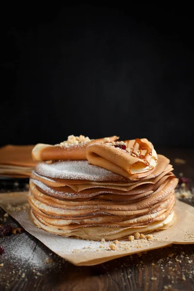 Dos pilas de panqueques, dos panqueques plegados, espolvoreados con bayas, galletas, azúcar en polvo y jarabe de caramelo en pergamino sobre una mesa de madera oscura — Foto de Stock