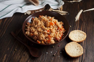 kahverengi bir seramik tabak, bir mutfak havlusu, kiraz domates ve bir sepetle spikelets buğday, ekmek ve koyu ahşap bir masa üzerinde bir tahta kaşık pilav