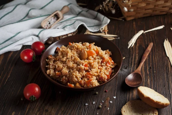 kahverengi bir seramik tabak, bir mutfak havlusu, kiraz domates ve bir sepetle spikelets buğday, ekmek ve koyu ahşap bir masa üzerinde bir tahta kaşık pilav