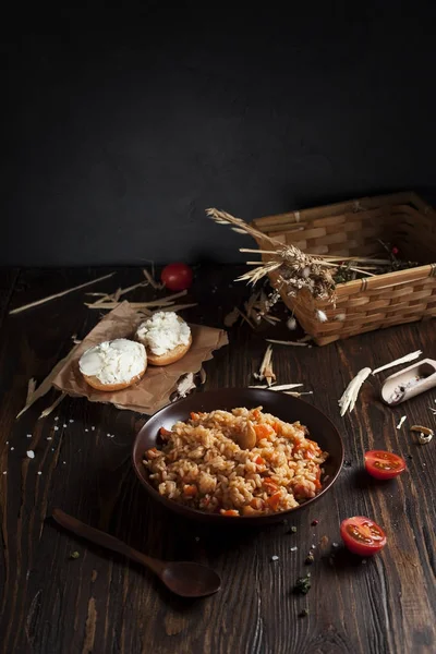 Плов в коричневой керамической тарелке, помидоры черри и корзина с шипами пшеницы, бутерброды с сыром и деревянная ложка на темном деревянном столе — стоковое фото