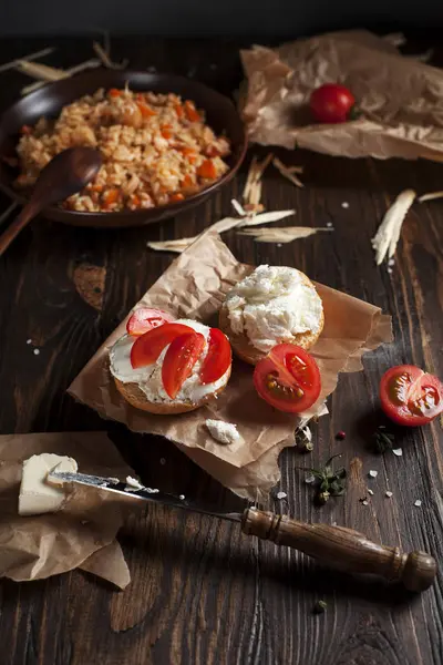 Тосты с сыром фета на выпечке бумаги, помидоры черри на фоне плова с овощами и курицей, на темном деревянном столе — стоковое фото