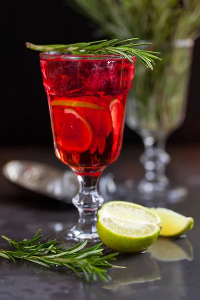 Una copa con un cóctel de color rojo con lima, romero y cubitos de hielo sobre un fondo de romero sobre un fondo oscuro — Foto de Stock