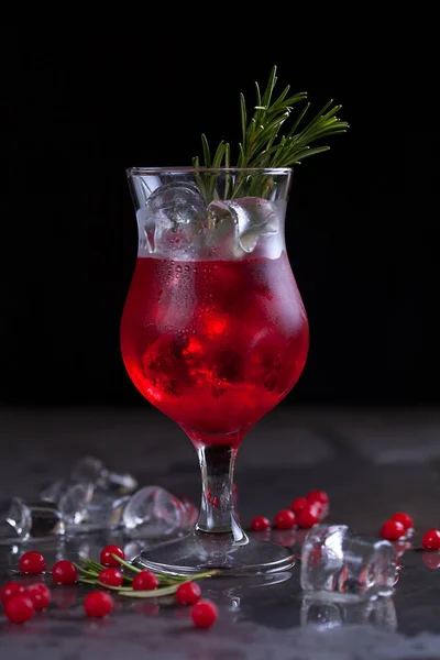 Un bicchiere con un cocktail di colore rosso con lime, bacche rosse, rosmarino e cubetti di ghiaccio su uno sfondo di rosmarino su sfondo scuro — Foto Stock