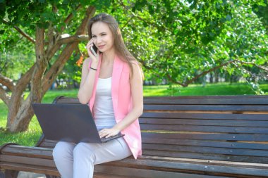 cep telefonuyla konuşurken ve dizüstü kullanarak parkta bankta oturan genç kadın.