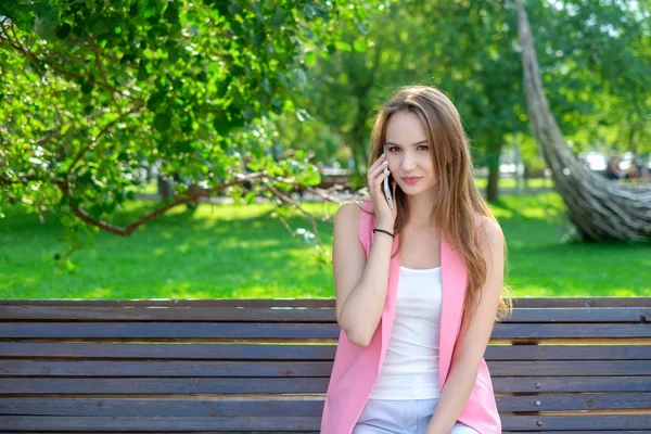 Portret van een aantrekkelijke jonge professionele vrouw met behulp van een smartphone zittend op een houten bank in een park, glimlachend. — Stockfoto