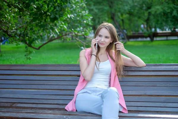 Portret van een aantrekkelijke jonge professionele vrouw met behulp van een smartphone zittend op een houten bank in een park, glimlachend. — Stockfoto