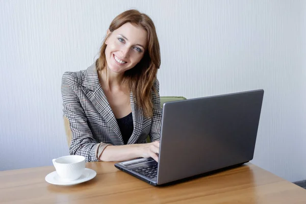 Χαριτωμένο θηλυκό νεαρός ενήλικας που εργάζονται στο φορητό υπολογιστή στο γραφείο δίπλα στο φλιτζάνι καφέ — Φωτογραφία Αρχείου