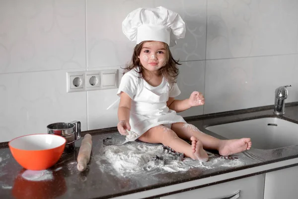 Милая маленькая девочка в фартуке и шляпе шеф-повара смешивает тесто и улыбается во время выпечки — стоковое фото