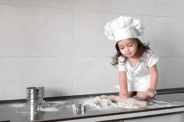 Sevimli küçük kız önlük ve Şef şapkalı hamur yoğurma ve pişirme sırasında gülümseyen — Stok fotoğraf