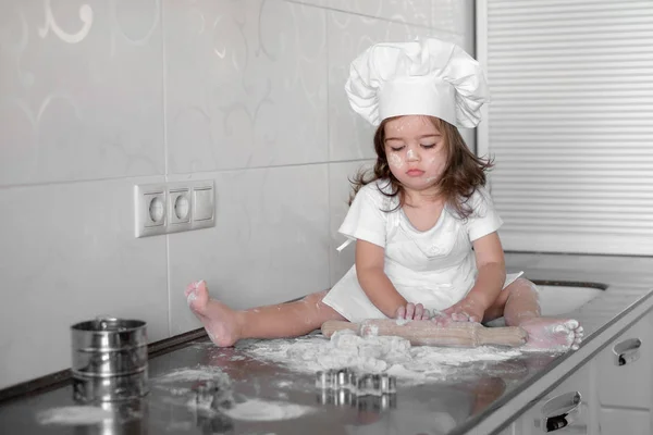Маленькая улыбающаяся маленькая девочка пекарь в белой шляпе повара и фартук разминает тесто на кухне — стоковое фото
