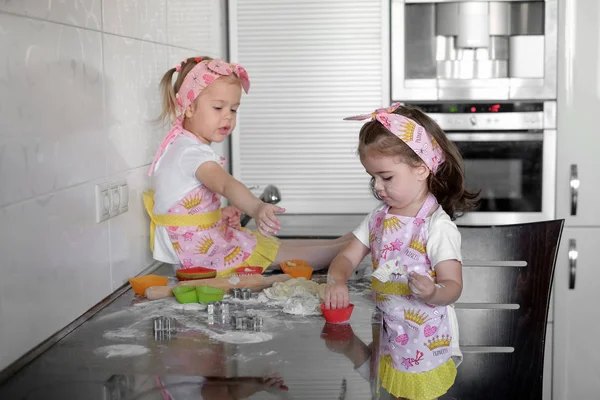 Two happy little girls child cook with flour and tough at the table in the kitchen is lovely and beautiful — стоковое фото