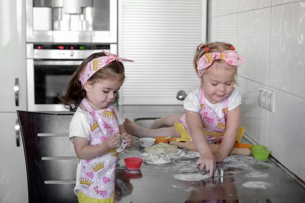 Two happy little girls child cook with flour and tough at the table in the kitchen is lovely and beautiful — стоковое фото