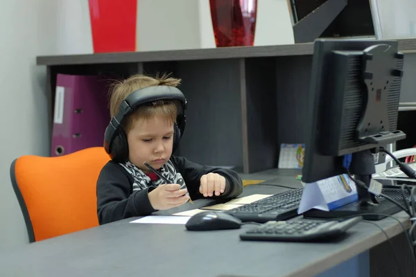 Jongetje met koptelefoon zit op computer in kantoor en maakt notities — Stockfoto