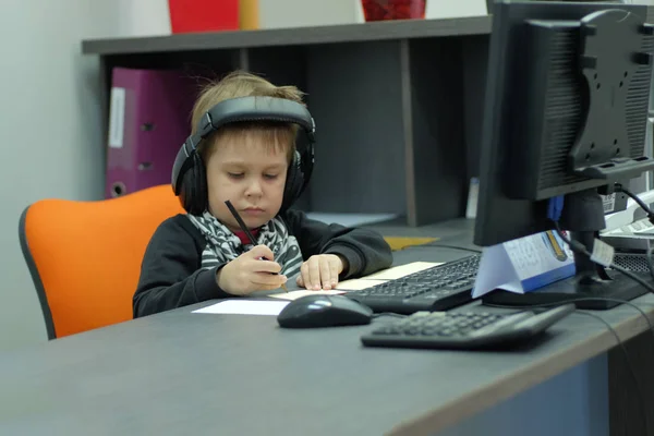 Niño pequeño con auriculares sentado en la computadora en la oficina y toma notas —  Fotos de Stock