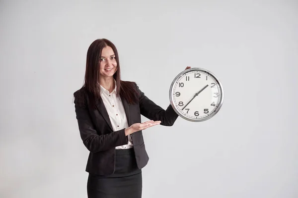 Деловая женщина показывает на большой круглые часы, на сером фоне стены в офисе Лицензионные Стоковые Изображения