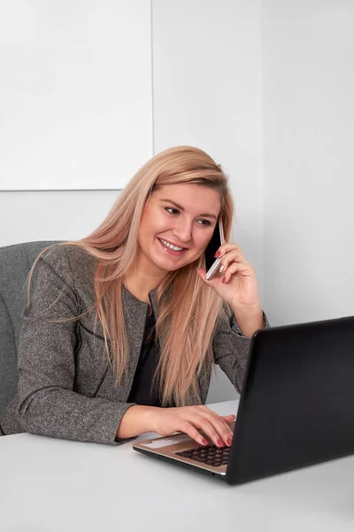 Gros plan d'une femme d'affaires intéressée dans un costume travaillant à son bureau blanc. Elle parle au téléphone et travaille avec un ordinateur portable — Photo