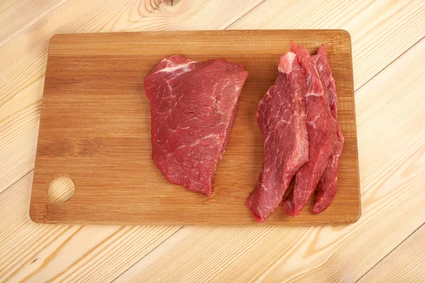 Peça de carne crua e fatias em tábua de corte de madeira — Fotografia de Stock