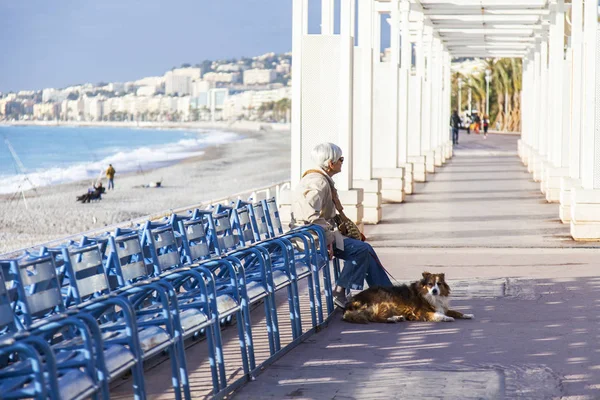 NICE, FRANCE, on JANUARY 6, 2017. The sun lights terraces on Promenade des Anglais - the main embankment of the city, one of the most beautiful in the world. The woman with a dog sits on a bench — Stock Photo, Image