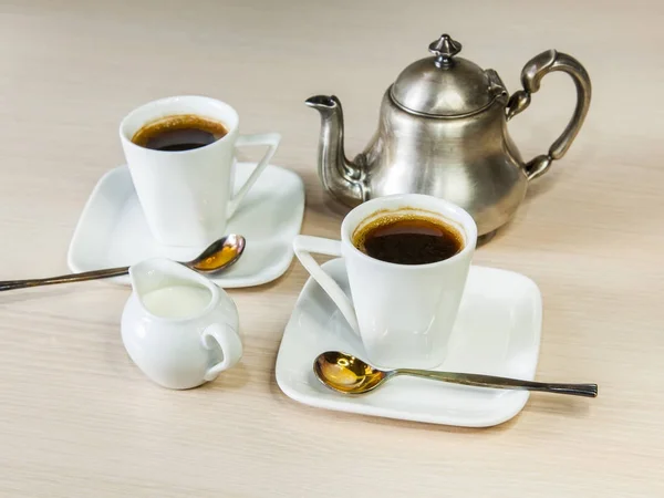 Two white porcelain cups with coffee of espresso and a silver coffee pot on a table