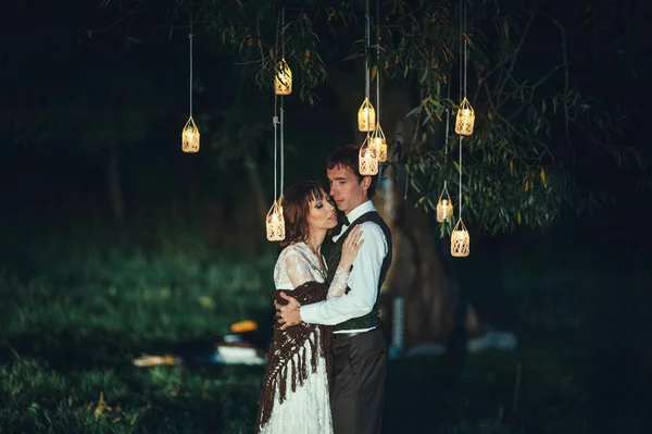 young beautiful engaged couple hugging beside tree with lanterns outdoors at nighttime