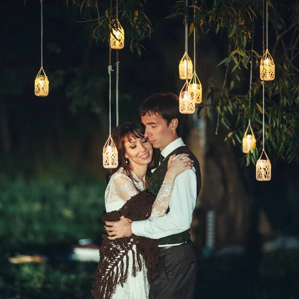 young beautiful caucasian engaged couple hugging under tree with lanterns outdoors at nighttime