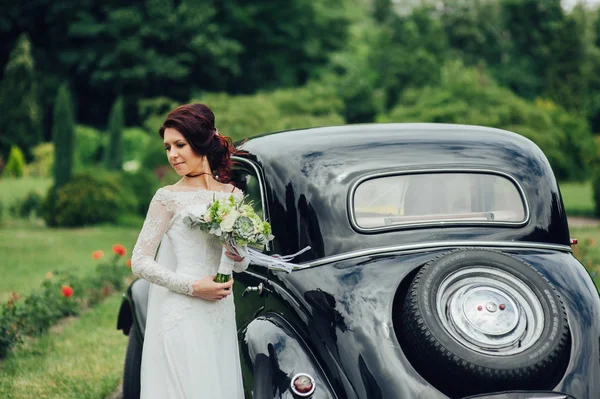 caucasian bride with flowers posing beside retro car in park