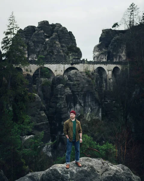 man standing on rock with mountains, forest and Bastei bridge in Saxon Switzerland on background