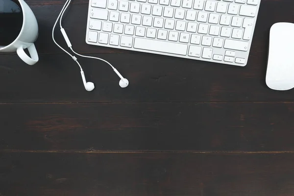 White computer keyboard mouse earphones and coffee cup overhead on dark brown table background