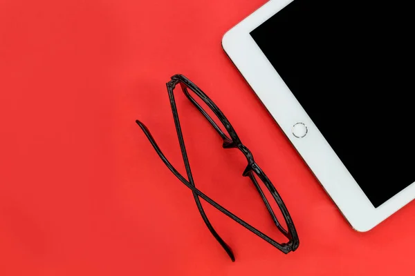 White tablet computer earphones and reading eyeglasses overhead on bright red background