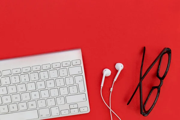 Computer keyboard white music earphones with eyeglasses on bright red background
