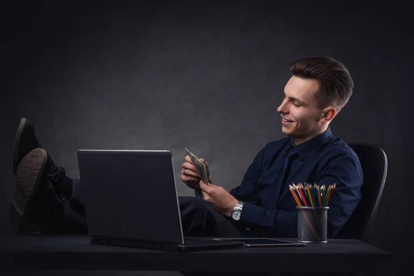 Lyckad affärsman vid bordet på en mörk bakgrund med bärbar dator och pengar. — Stockfoto