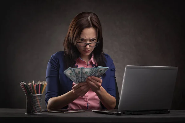 Traurige Geschäftsfrau am Tisch vor dunklem Hintergrund mit Laptop und Geld. — Stockfoto